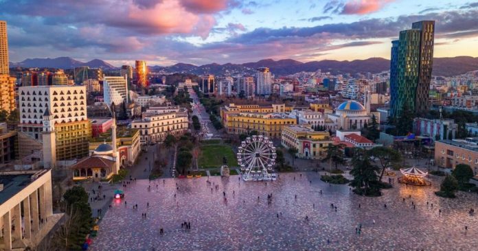 An Albanian city square with a Ferris wheel and mountains beneath a pink sky.