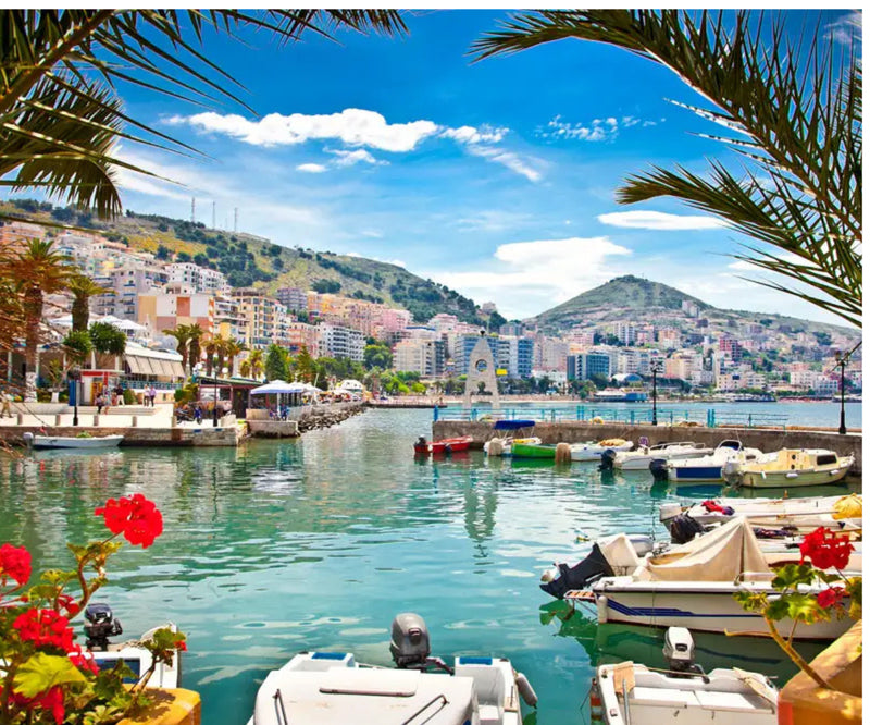 Small boats in an Albanian harbor framed by palms, flowers and hillside buildings.