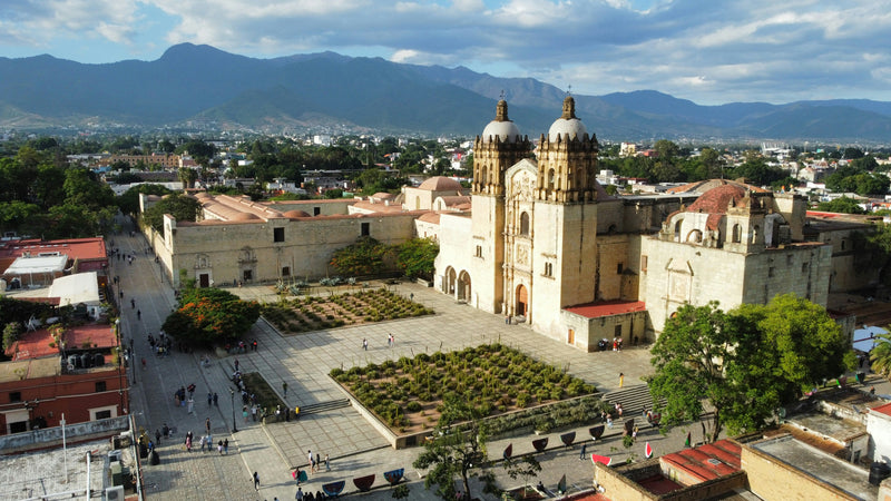 A stone church and plaza amid Oaxaca rooftops with mountains beyond.